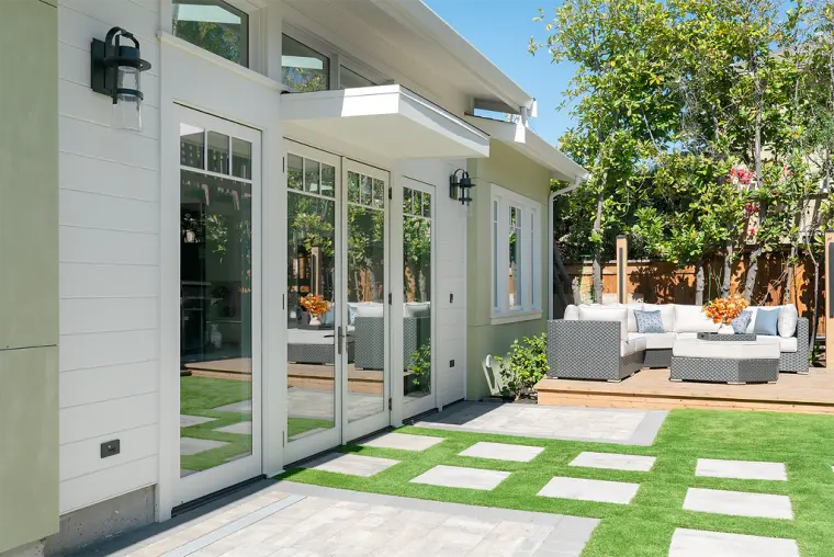 French doors along the white lap-sided wall, opening onto a stepping-pad lawn and a deck with an outdoor sectional
