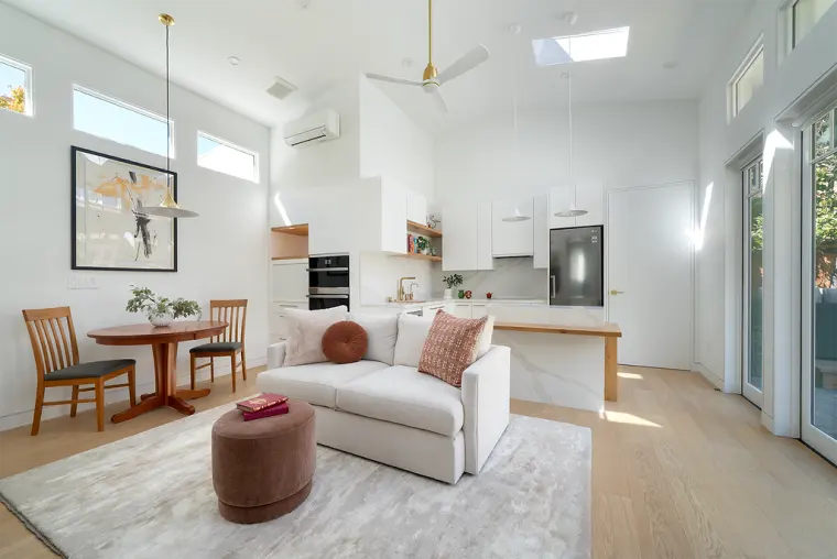 Open great room under a vaulted ceiling with a skylight, a cream loveseat, a round dining table, and a white kitchen with a waterfall island