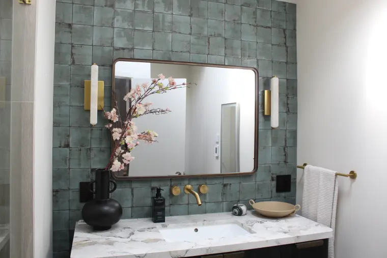 Bathroom vanity wall with green handmade square tile, a rounded dark-framed mirror, brass wall faucet and sconces, and a marble counter