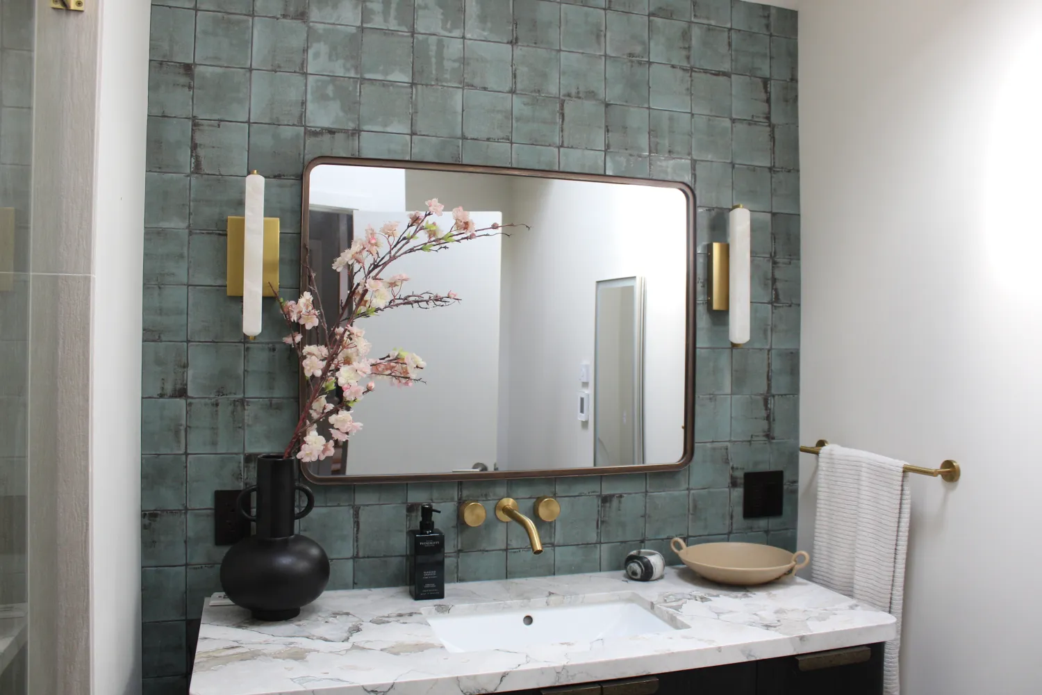 Bathroom vanity wall with green handmade square tile, a rounded dark-framed mirror, brass wall faucet and sconces, and a marble counter