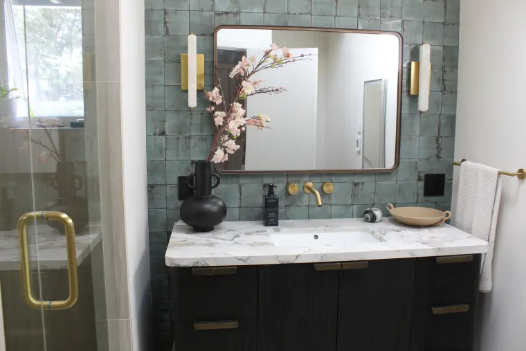 Dark wood vanity with a marble top and green tile wall beside a frameless glass shower door with a brass handle