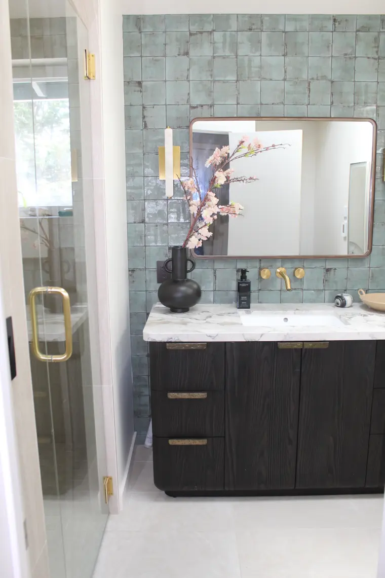 Vertical view of the dark wood vanity, green tile wall, and mirror next to a glass shower door with a brass pull
