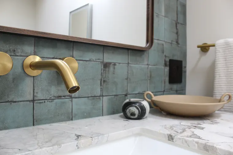 Brushed brass wall-mounted faucet over an undermount sink with a marble counter and green glazed tile backsplash