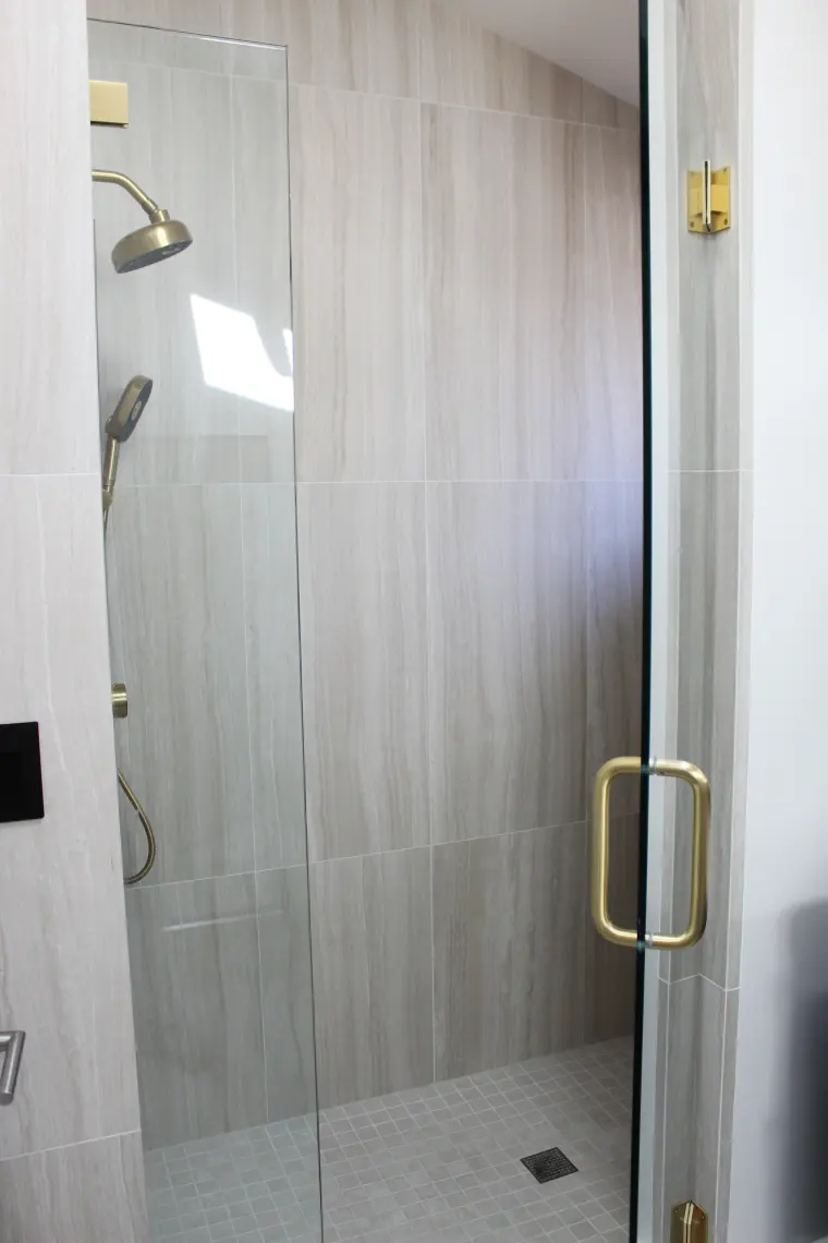 Frameless glass shower door with a brass pull opening to a shower with wood-look wall tile and a small-tile floor