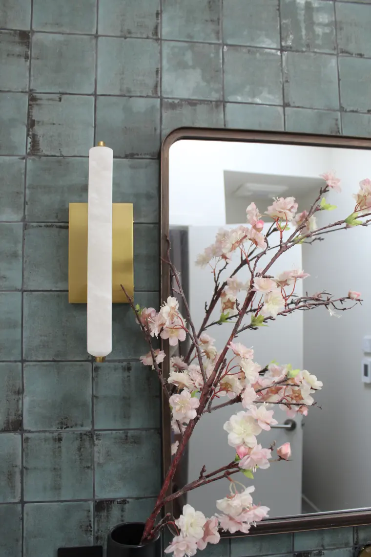 Detail of a brass and alabaster sconce beside a dark-framed mirror on green glazed tile, with cherry blossom stems