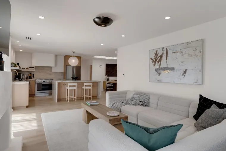 Open living room with a white sectional and coffee table looking toward a kitchen with light wood cabinets, island stools, and a globe pendant
