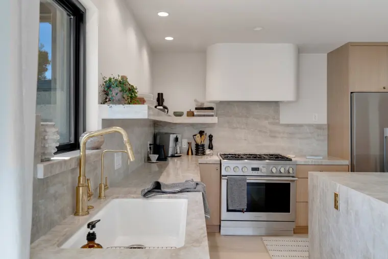 Kitchen sink wall with a white farmhouse sink, brass faucet, marble backsplash, open shelves, and a stainless gas range under a white hood