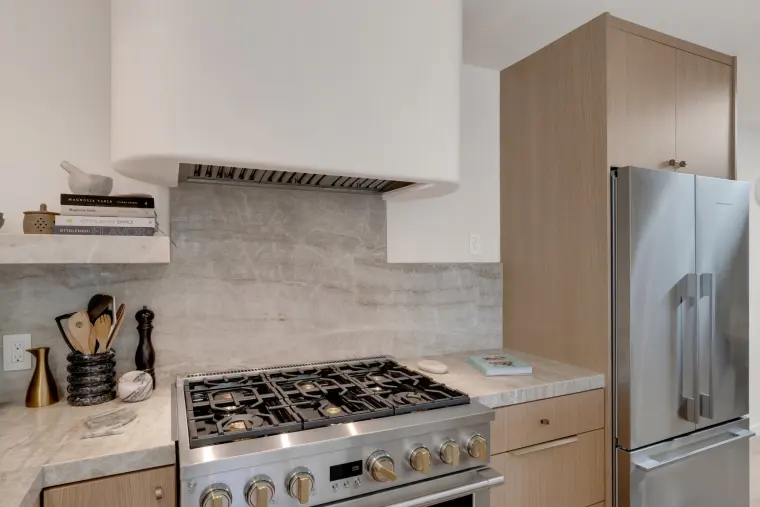 Stainless gas range and French-door refrigerator beside light wood cabinets with a marble backsplash and white plaster hood
