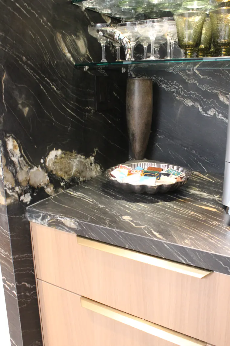 Bar counter in black and gold veined stone with a full-height stone backsplash, glass shelves, and light wood drawers with brass pulls