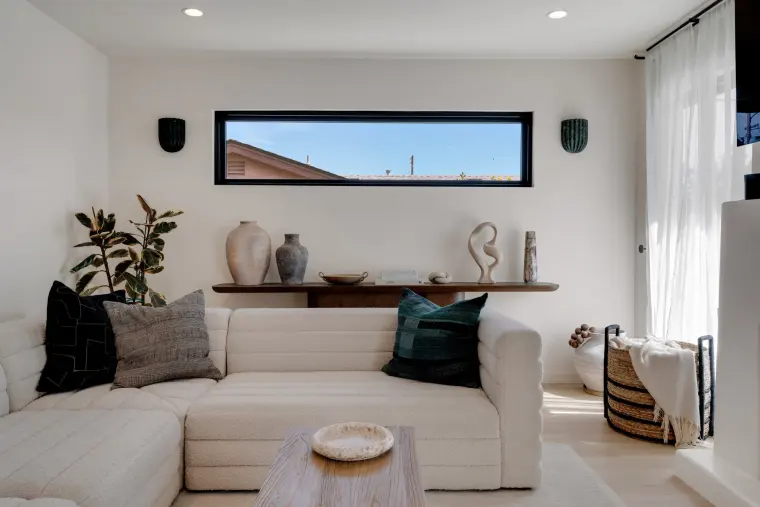 White sectional sofa under a long clerestory window with a floating wood shelf holding vases and sculptures