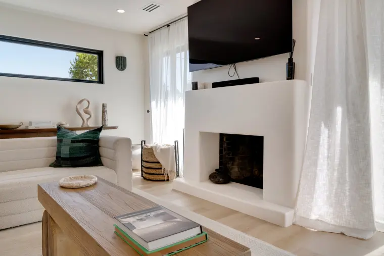 White plaster fireplace with a wall-mounted TV above, a clerestory window, and a wood coffee table with books