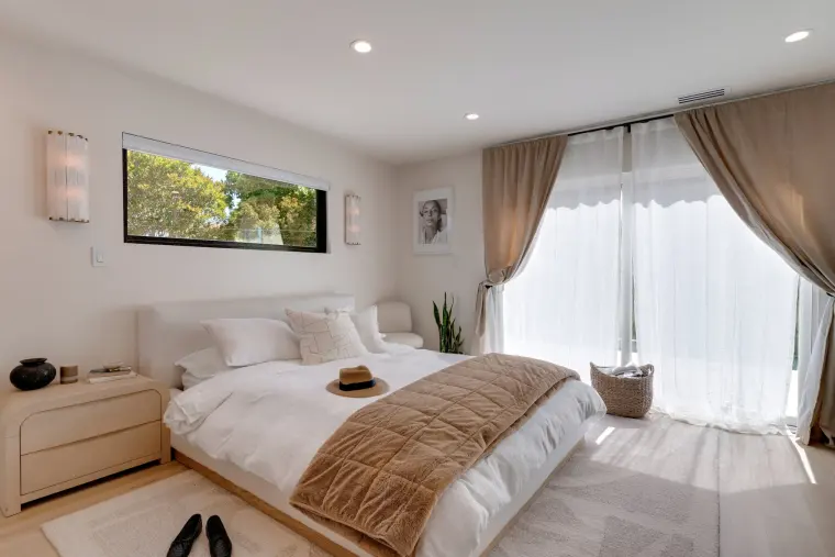 Bedroom with a white upholstered bed, clerestory window flanked by sconces, and sheer and taupe curtains over a sliding door