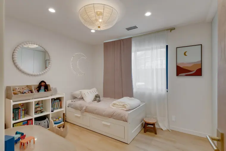 Child bedroom with a white daybed, low bookshelves, a round mirror, flush ceiling light, and a window with sheer curtains