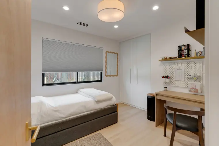 Small bedroom with a daybed, wall desk with a wooden chair, floor-to-ceiling white closet doors, and a window with a shade