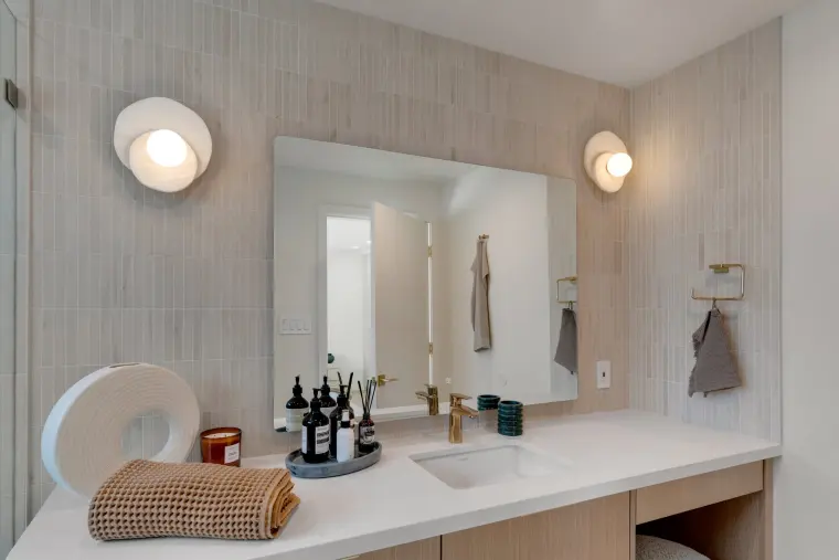 Vanity with a white counter, light wood drawers, frameless mirror, round plaster sconces, and vertical stacked tile wall