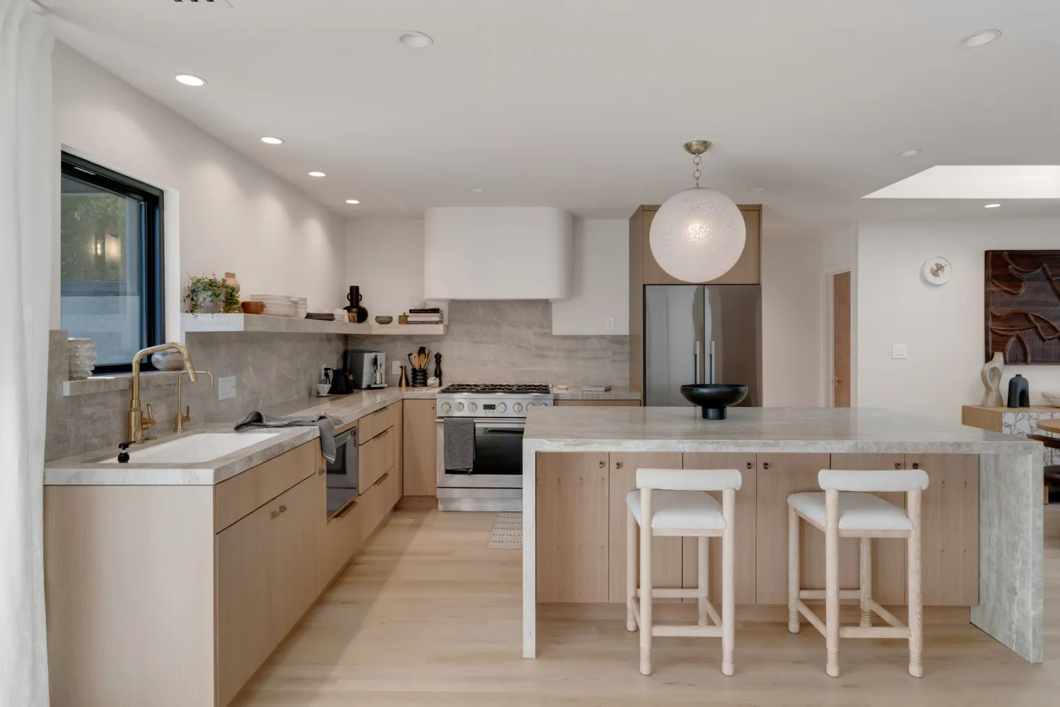 Kitchen with pale oak lower cabinets, a white plaster hood, stone island with counter stools, and a skylight