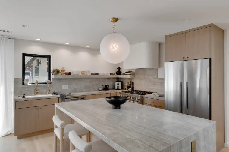 Stone kitchen island in front of pale oak cabinetry and a stainless refrigerator