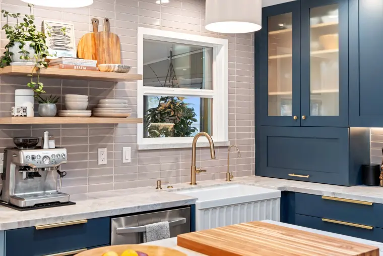 Farmhouse sink with a brass faucet under open wood shelves, gray tile backsplash, and navy glass-front cabinets