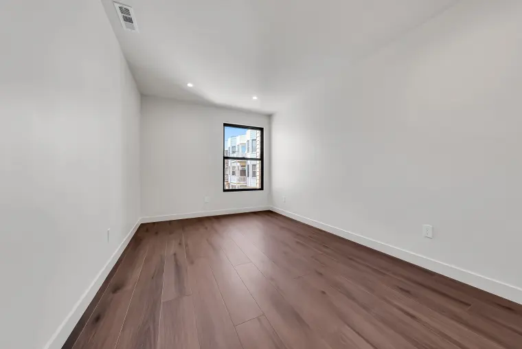Bedroom corner with a tall black-framed window overlooking neighboring rowhouses and wood flooring