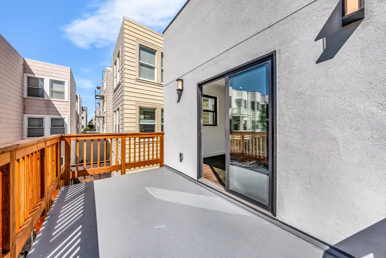 Rear wood deck with stained railings beside a white stucco wall and black-framed sliding glass door, neighboring rowhouses behind