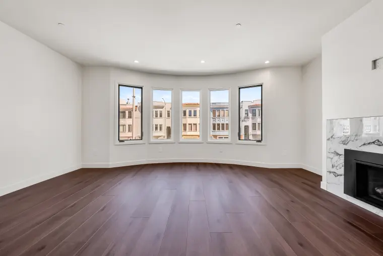 Empty living room with a curved five-window bay, wide-plank wood flooring, and the edge of a marble fireplace surround