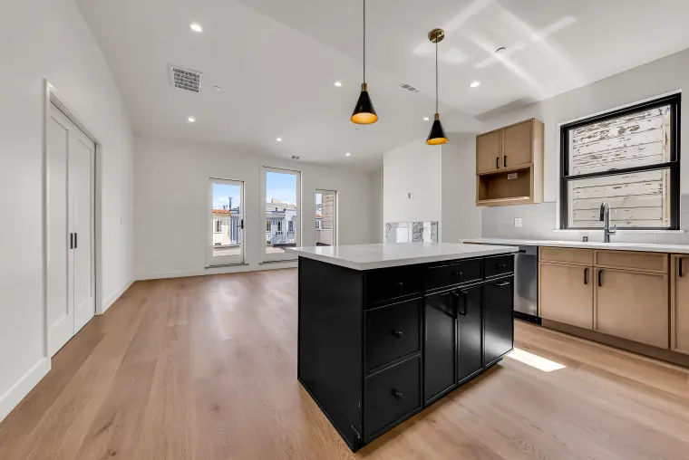 Kitchen with a black island, light oak cabinets, black cone pendants, and a window over the sink, opening to glass doors beyond
