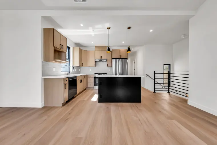 Kitchen with light oak cabinets, a black island, stainless appliances, black cone pendants, and a black horizontal stair railing