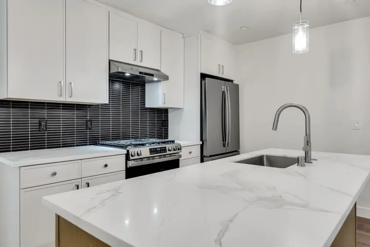 Kitchen with white shaker cabinets, black stacked tile backsplash, a gas range, stainless refrigerator, and a veined quartz island with sink