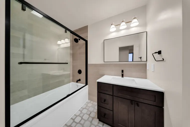 Bathroom with a glass tub enclosure, a dark wood vanity with white top, a black-framed mirror, and patterned floor tile