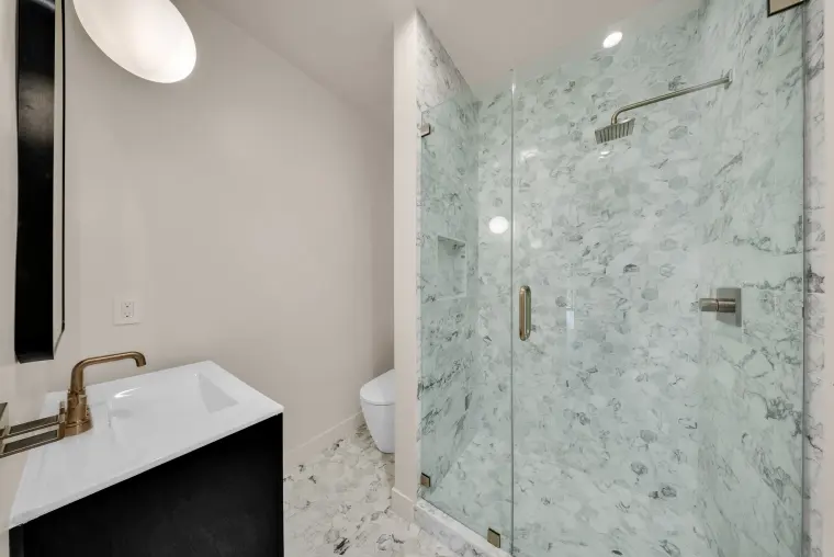 Bathroom with a frameless glass shower in marble-look tile, a black vanity with brass faucet, and a globe wall light