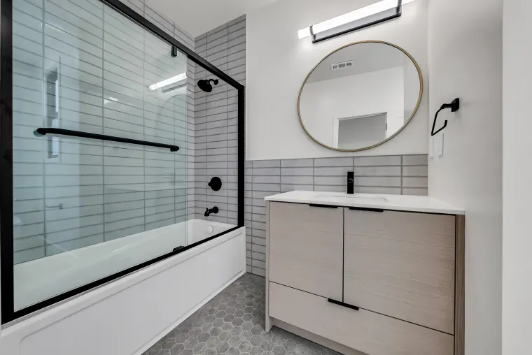 Bathroom with a black-framed glass tub enclosure, gray tile walls, a light oak vanity, and a round brass mirror