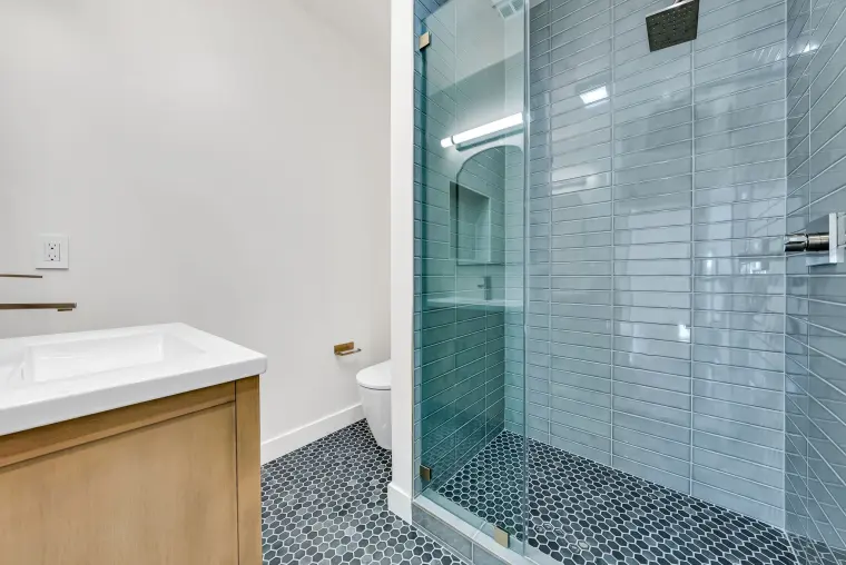 Walk-in shower with blue-gray stacked glass tile, a frameless glass door, a mosaic floor, and a light oak vanity