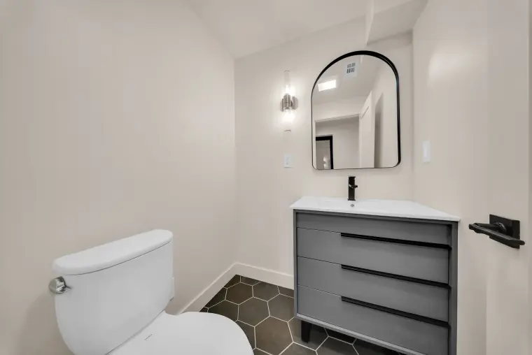 Bathroom with a gray drawer vanity, arched black-framed mirror, wall sconce, and black hexagon floor tile
