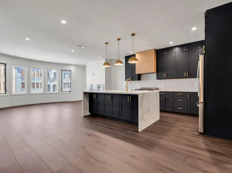 Finished open-concept kitchen with matte black shaker cabinets, brass pendant lights, waterfall quartz island, and wide-plank flooring opening to an empty bay-window living area