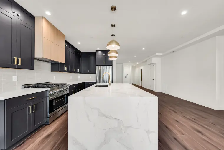 Kitchen with black shaker cabinets, a light wood range hood, brass pendants, and a waterfall quartz island looking down a long hallway