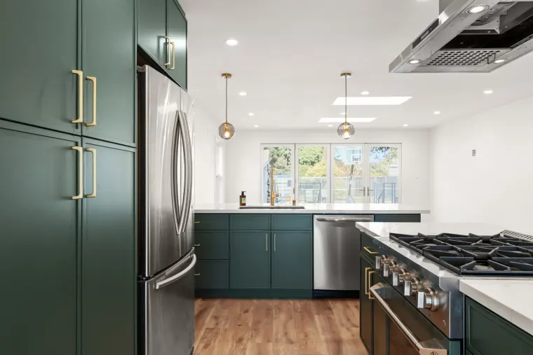 Green cabinets with brass pulls, stainless refrigerator, and smoked glass pendants over the island