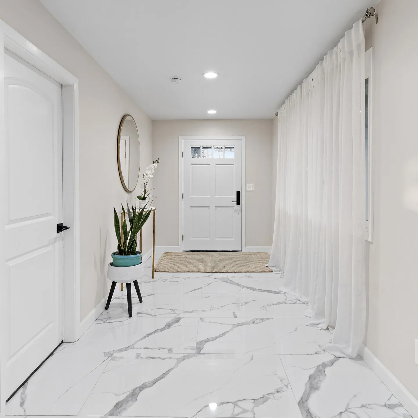 Finished entry hallway with polished marble-look porcelain floor tile, sheer floor-to-ceiling curtains, round brass mirror, console table with orchid and snake plant, white paneled front door with jute welcome mat