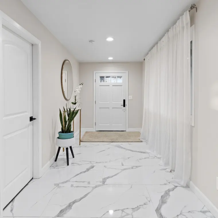 Finished entry hallway with polished marble-look porcelain floor tile, sheer floor-to-ceiling curtains, round brass mirror, console table with orchid and snake plant, white paneled front door with jute welcome mat