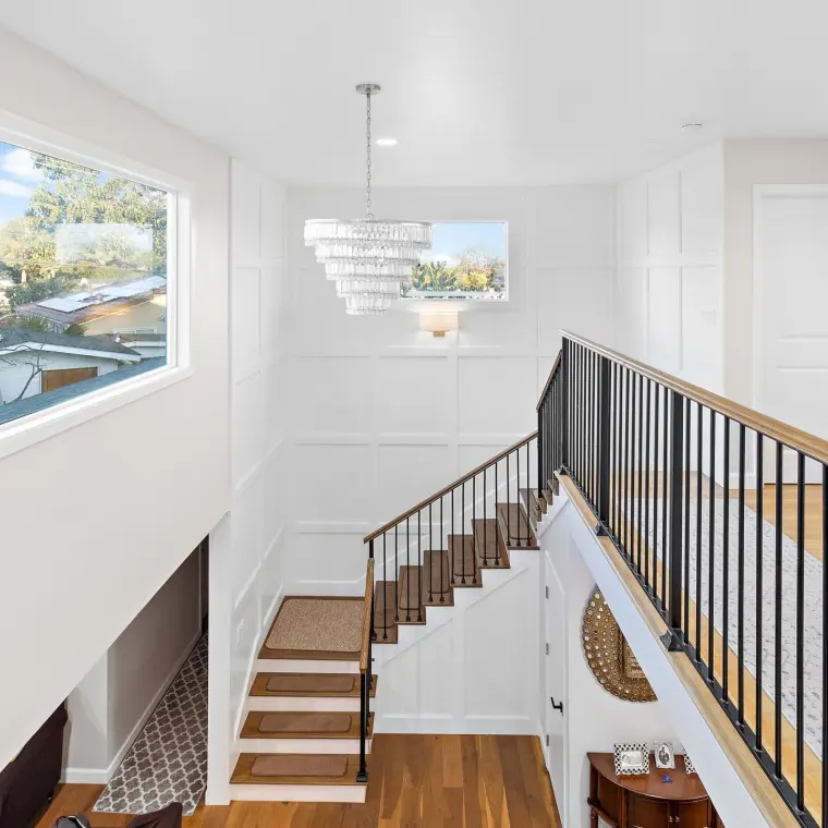 Two-story stairwell/foyer viewed from the upper landing, tiered crystal chandelier, tall wainscoted walls, window with neighborhood view, decorative round mirror and console table visible below