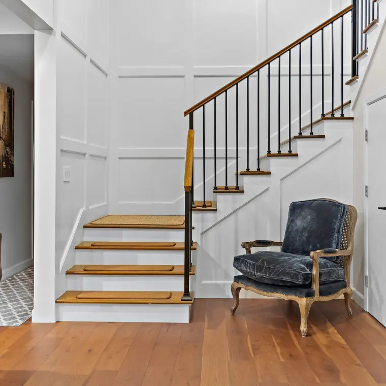 Finished staircase with full-height wall paneling/wainscoting, wood handrail with black iron balusters, sisal stair runners on wood treads, a blue velvet French bergere chair, wide-plank walnut floors, framed street photograph on the wall