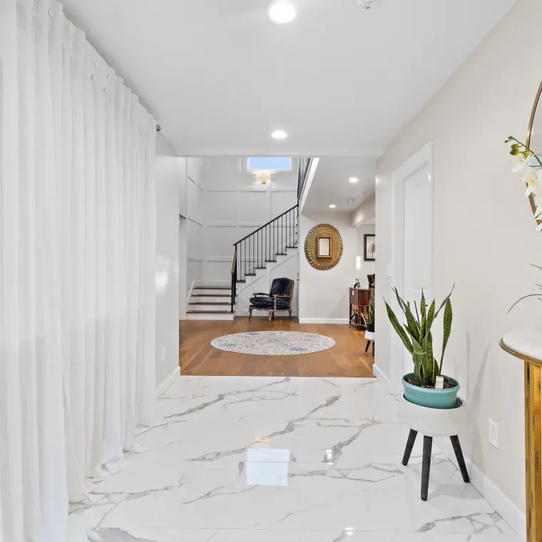 View from the entry hall into the finished stairwell/foyer, blue velvet bergere chair on a round patterned rug, decorative wall mirror, wood floors transitioning to marble-look tile in the foreground