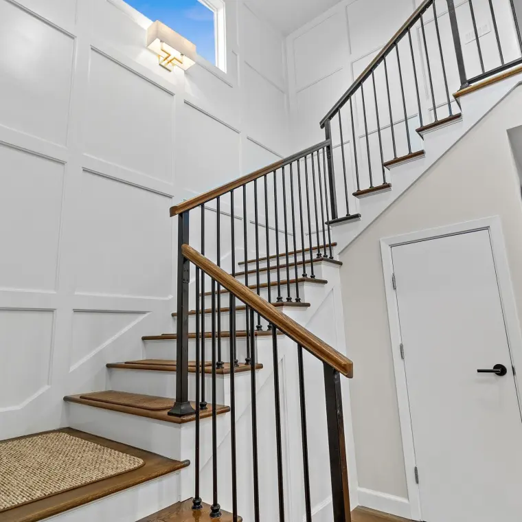 Two-story stairwell with full-height wall paneling, wood handrails with black iron balusters, sisal stair runners, a brass sconce beneath a small window, white paneled door to a closet below the stairs