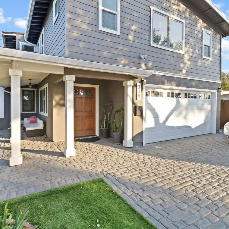 Finished two-story craftsman-style home exterior, gray horizontal siding, white porch columns and trim, wood front door, two-car garage, cobblestone-pattern driveway and new artificial turf strip