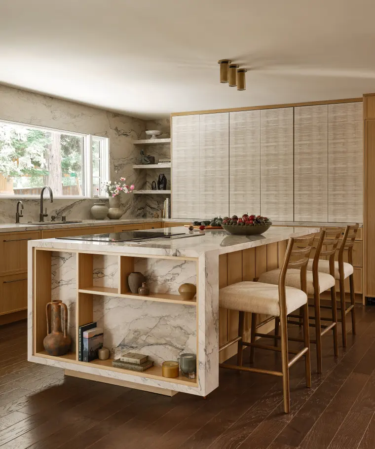 Kitchen island wrapped in veined stone with open oak display shelves, upholstered counter stools, and textured pantry doors behind