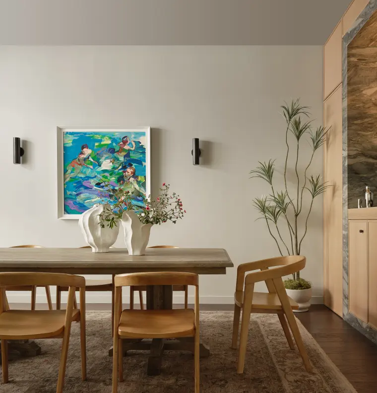 Dining room with a wood trestle table, oak chairs, white sculptural vases, a colorful painting between black sconces, and a tall potted plant