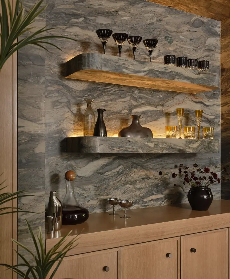 Angled view of the built-in bar with lit floating stone shelves holding glassware and decanters above white oak cabinets