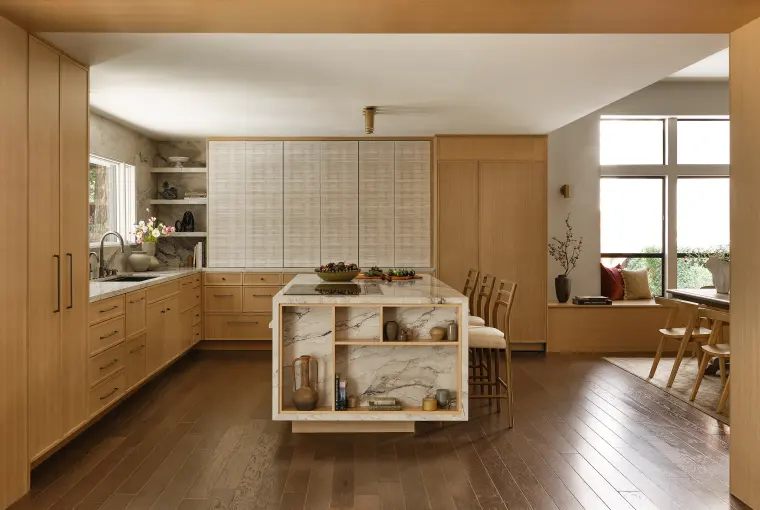 The kitchen from the living side: white oak cabinets, stone island with display shelving, and a window seat beyond