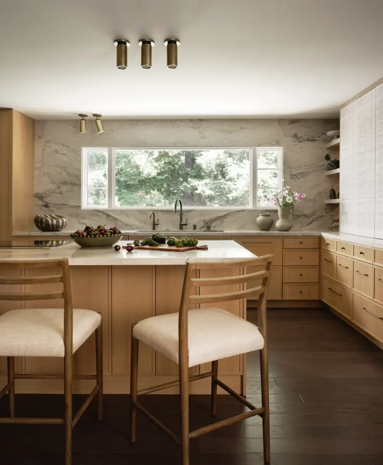 Stone island with cushioned counter stools under brass ceiling lights, the sink window behind